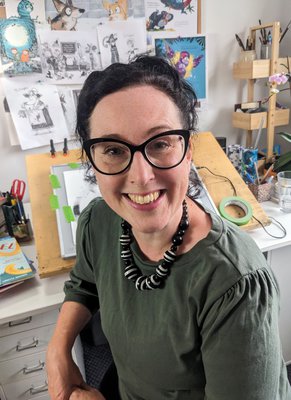 A photograph of children's author Loretta Schauer - a smiling white woman with black hair, tied back, and black framed cats-eye glasses. Behind her is a drawing board and pinned up examples of her illustrations.