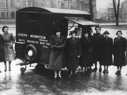 Black and White Image of women in coats and hats stood in front of a trailer.