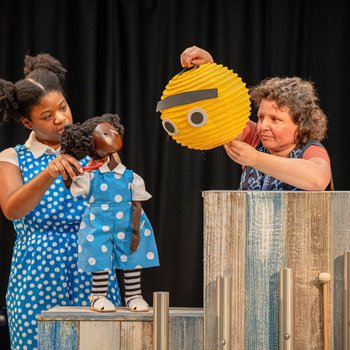 Puppet Ruby stands on a stack of boxes, looking at a small, yellow worry puppet