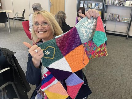 A woman with blonde hair sat holding a care squares blanket with colourful knitted squares