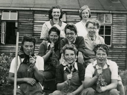 A group of women from the Land Army standing in front of a hut, holding carrots.