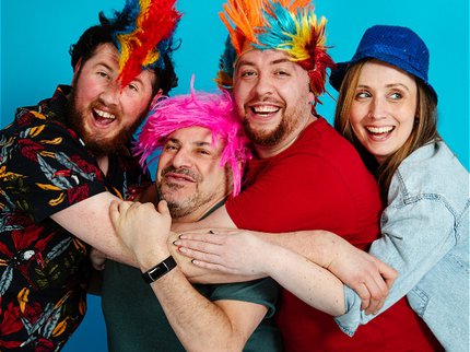 Four people wearing colourful feathers stood against a blue background.