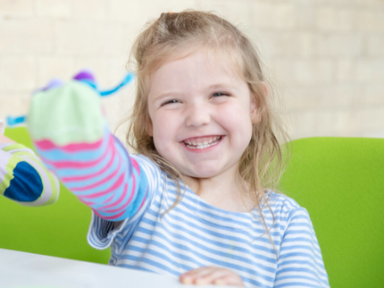 A child with short blonde hair holds up a blue sock puppet and smiles