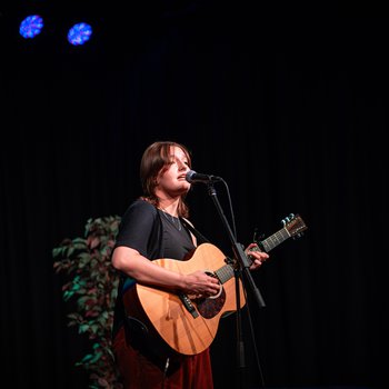 A girl with short brown hair wearing a black t shirt playing the acoustic guitar and singing.