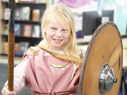 A girl with blonde hair, dressed in a pink Viking tunic and holding a spear and shield