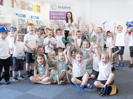 A group of children sat down in the library with their arms raised up and smiling, with an author holding up her book