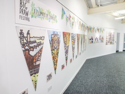 Wall in library gallery showing colourful drawings on bunting and photographs of the artist.