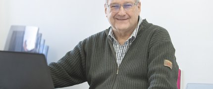 A white man with grey hair wearing glasses and smiling sits at a desk with a laptop ready to learn.