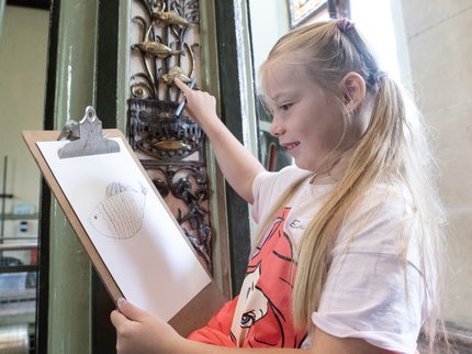 A girl in a white tshirt and pink dress sits with a clipboard and draws