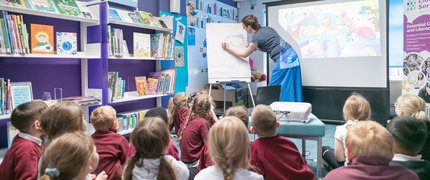 An author writes on a white board in front of a class of children sat in the library