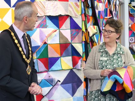 A woman smiles and holds a knitted blanket while talking to a man