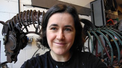 Photo of a women with short dark hair stood in front of a sculpture of a horse made from recycled metal pieces