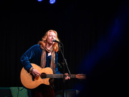 A man with long red hair playing the acoustic guitar and singing. He is wearing a blue shirt and orange patterned scarf.