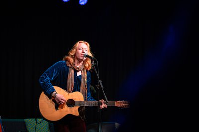 A man with long red hair playing the acoustic guitar and singing. He is wearing a blue shirt and orange patterned scarf.