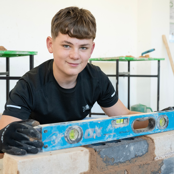 A construction student smiles at the camera whilst levelling a brick wall