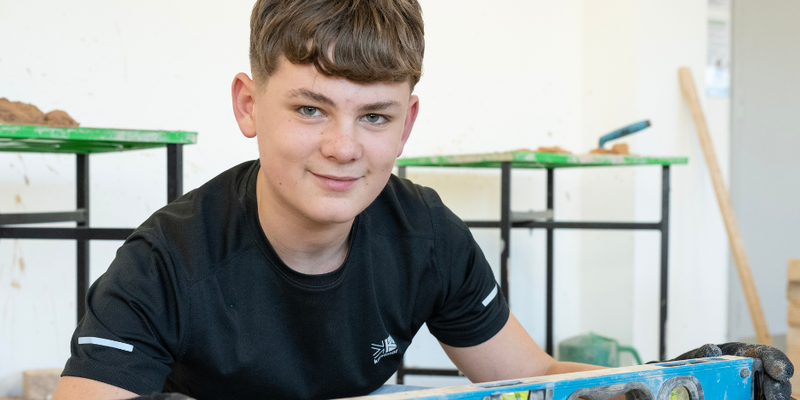 A construction student smiles at the camera whilst checking that a brick wall is level