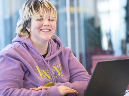 A student in a light purple hoodie is using their laptop and smiling at the camera