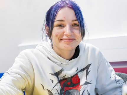 A young feminine-presenting person with purple hair in a ponytail is smiling at the camera