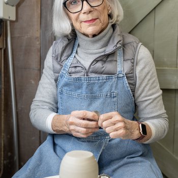 Photo of woman sat behind a potters wheel with a upturned pot on the wheel head.