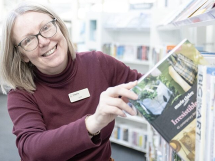 A woman taking a book from a library shelf and smiling at the camera