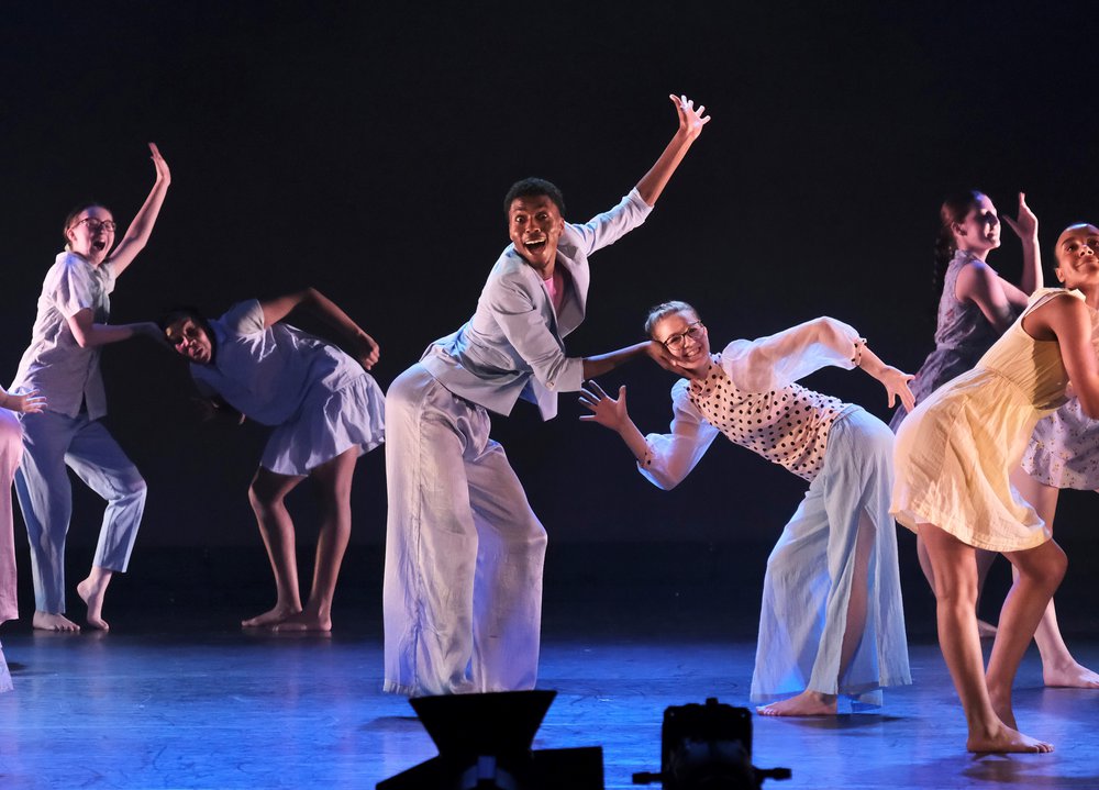 6 dancers on stage in pairs, wearing light costumes. half of the dancers have a hand in the air waving, the other holding their partners head who is bent over looking to the front smiling with both arms bent at the elbow.