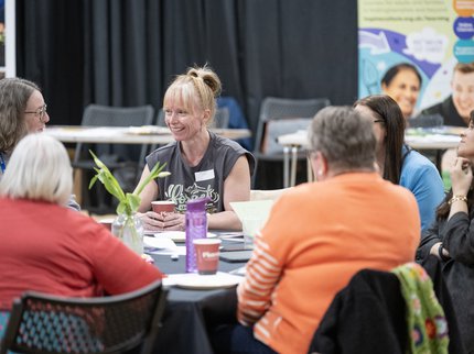 A woman smiling in a grey t-shirt sat at a conference table