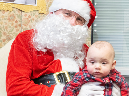 Santa is sitting in an ornate chair in his grotto! A baby is on his knee