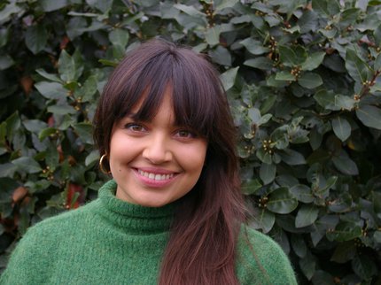 Photo of author Sara Nisha Adams - a woman of Indian-Kenyan and English descent, with long dark hair, wearing a green jumper.