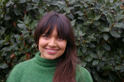 Photo of author Sara Nisha Adams - a woman of Indian-Kenyan and English descent, with long dark hair, wearing a green jumper.