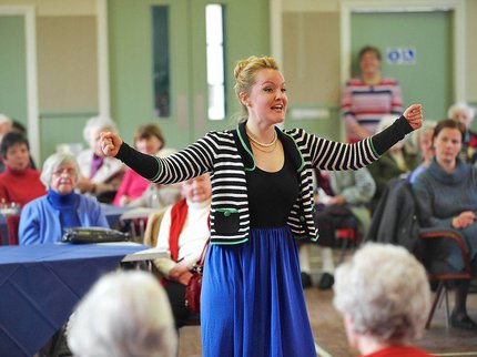 A woman in a stripy black and white cardigan and a blue skirt raises her arms to the sides and sings