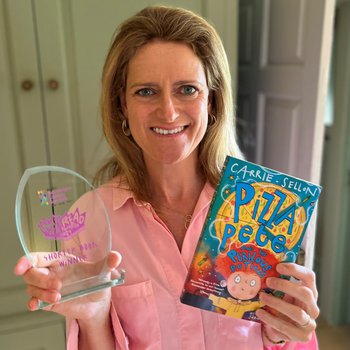 A woman with blonde hair and a pink shirt holds a book and a trophy, smiles at the camera