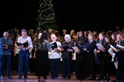 A group of adults singing in a semi-circle on stage with a christmas tree