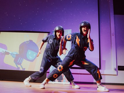 Two dancers with disco style helmets and purple jumpsuits strike a pose