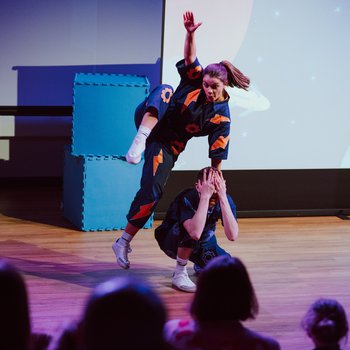 One dancer crouched with hands covering their eyes, while another leans one handed on his head, raising knee high in dance move.