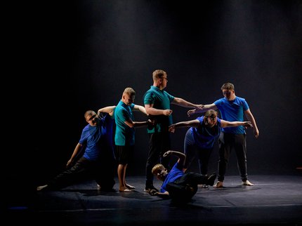 Six dancers on a black stage. Two in green t-shirts and four in blue t-shirts. One is on the floor, on his back with legs and arms in the air. A female is bent forward with her arms out to the side with the four other male dancers connected in a line