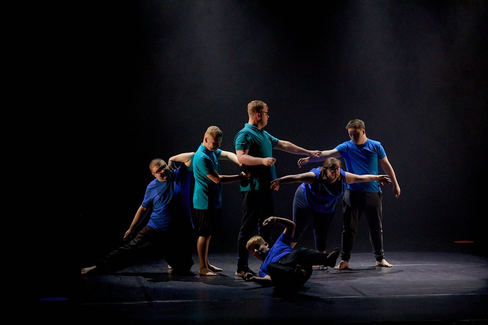 Six dancers on a black stage. Two in green t-shirts and four in blue t-shirts. One is on the floor, on his back with legs and arms in the air. A female is bent forward with her arms out to the side with the four other male dancers connected in a line