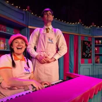 women in pink hat sat at piano, man in apron stood behind her.