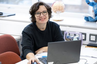 Woman aged 30-40 with short dark wavy hair and glasses sits at a computer desk smiling.