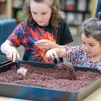 Two girls digging for artefacts in a small soil tray.