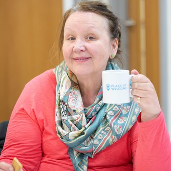 Smiling lady holding in bright jumper and scarf holding her Places of Welcome mug