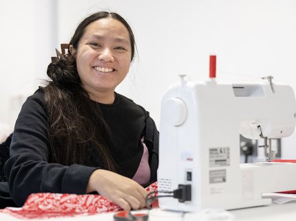 Smiling learner with dark hair wearing a black long sleeve t-shirt sits at a sewing machine using red patterned fabric and red cotton.